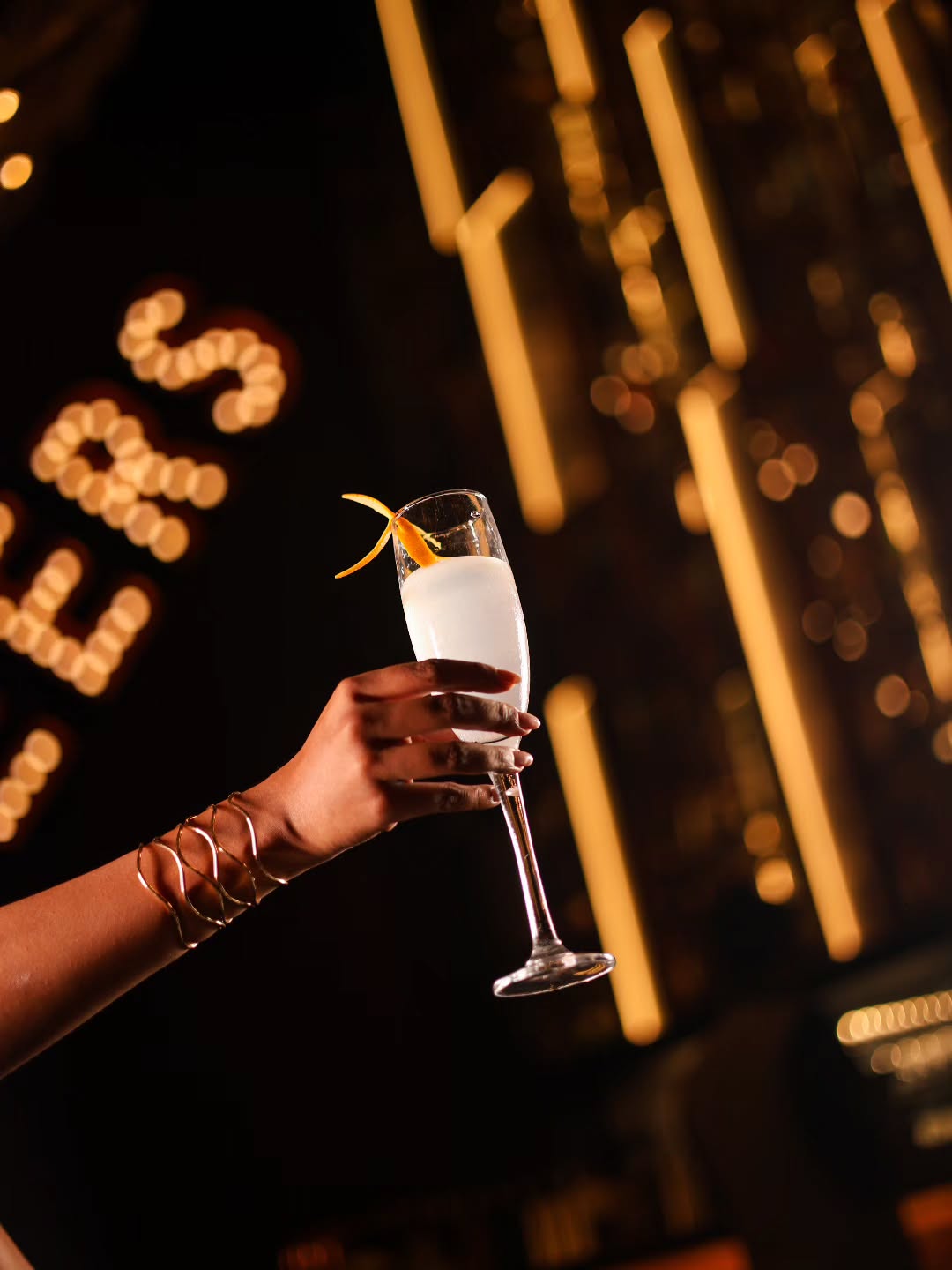 A signature cocktail with an orange twist raised against Arena's glowing Cheers sign